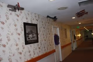 A hallway in a senior living facility with floral wallpaper, a wooden handrail, and several doors. The hallway is decorated with framed pictures and model airplanes hanging from the ceiling. The lighting is soft, and the corridor extends into the distance.