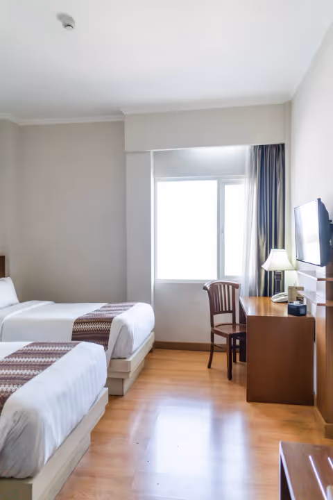Bright twin-bed bedroom with wooden floors, a desk and chair by a window, and a wall-mounted TV.