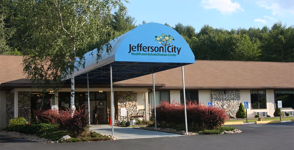 Front exterior view of Jefferson City Health and Rehabilitation Center building with a blue awning displaying the facility's name and logo, surrounded by trees and bushes under a partly cloudy sky.
