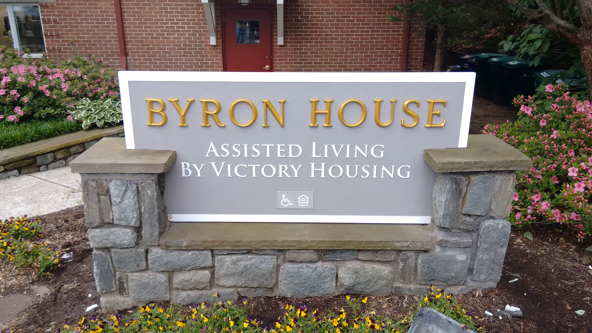 Stone and concrete sign for Byron House Assisted Living by Victory Housing, surrounded by flowers and greenery, with a brick building and a red door in the background.