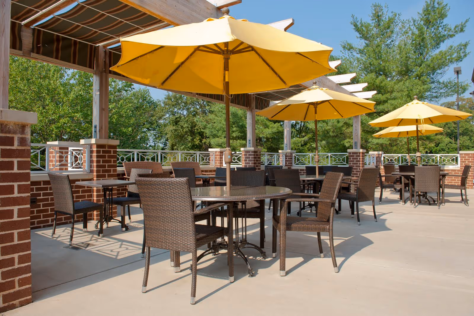 Outdoor patio area with several round tables and brown wicker chairs under large yellow umbrellas. The patio is surrounded by brick pillars and a metal railing, with green trees visible in the background under a clear blue sky.