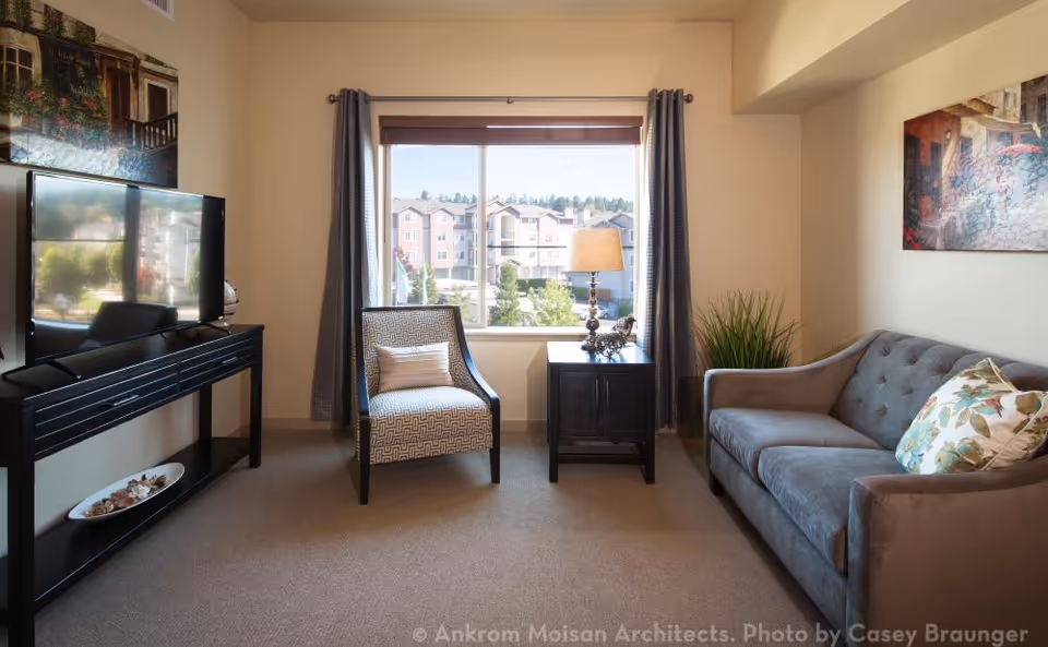 A furnished living room with a sofa, armchair, TV console and a window overlooking nearby apartment buildings.