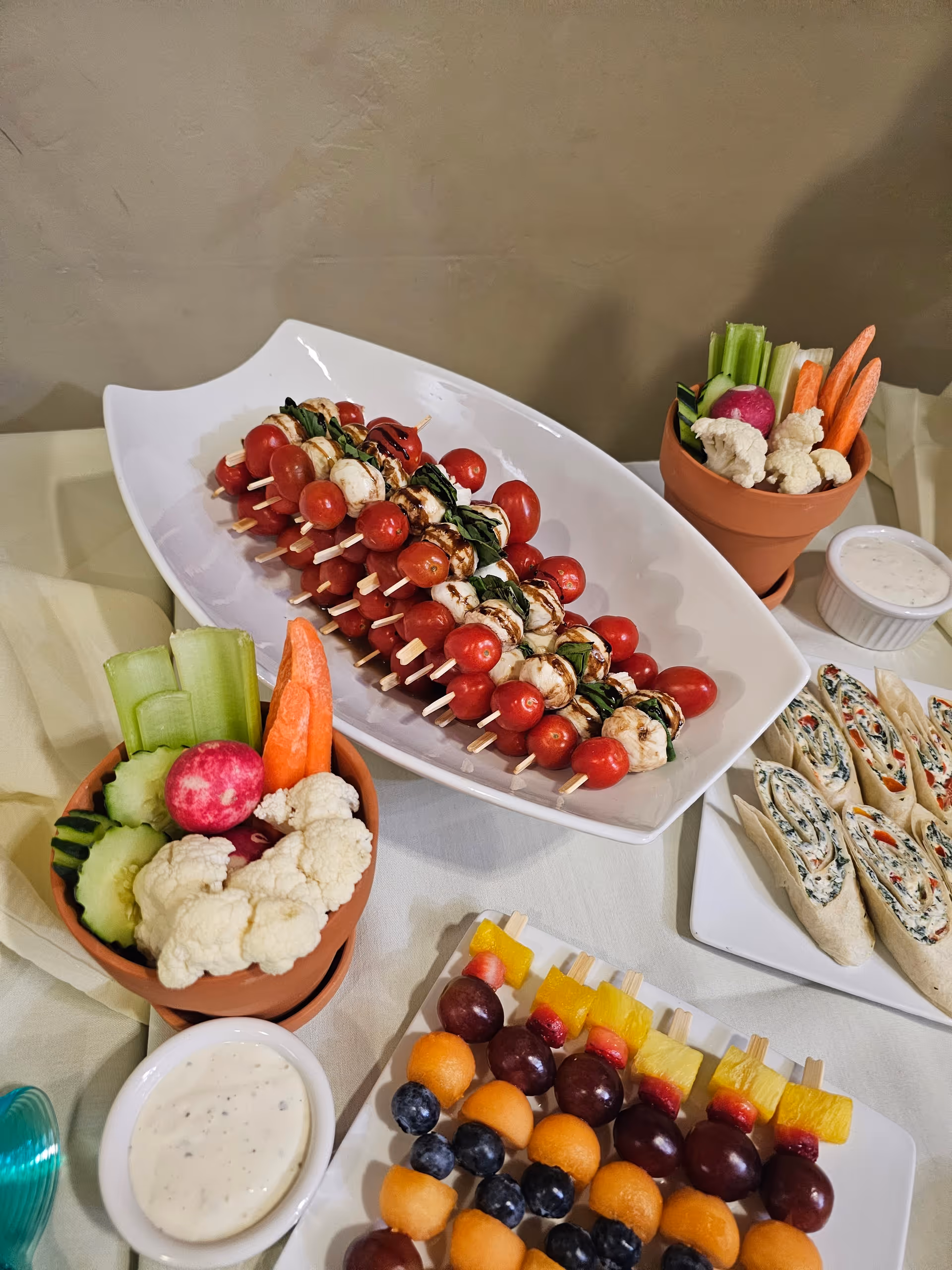 Platter of skewered tomatoes, mozzarella and basil with vegetable cups, fruit skewers and dips on a buffet table.