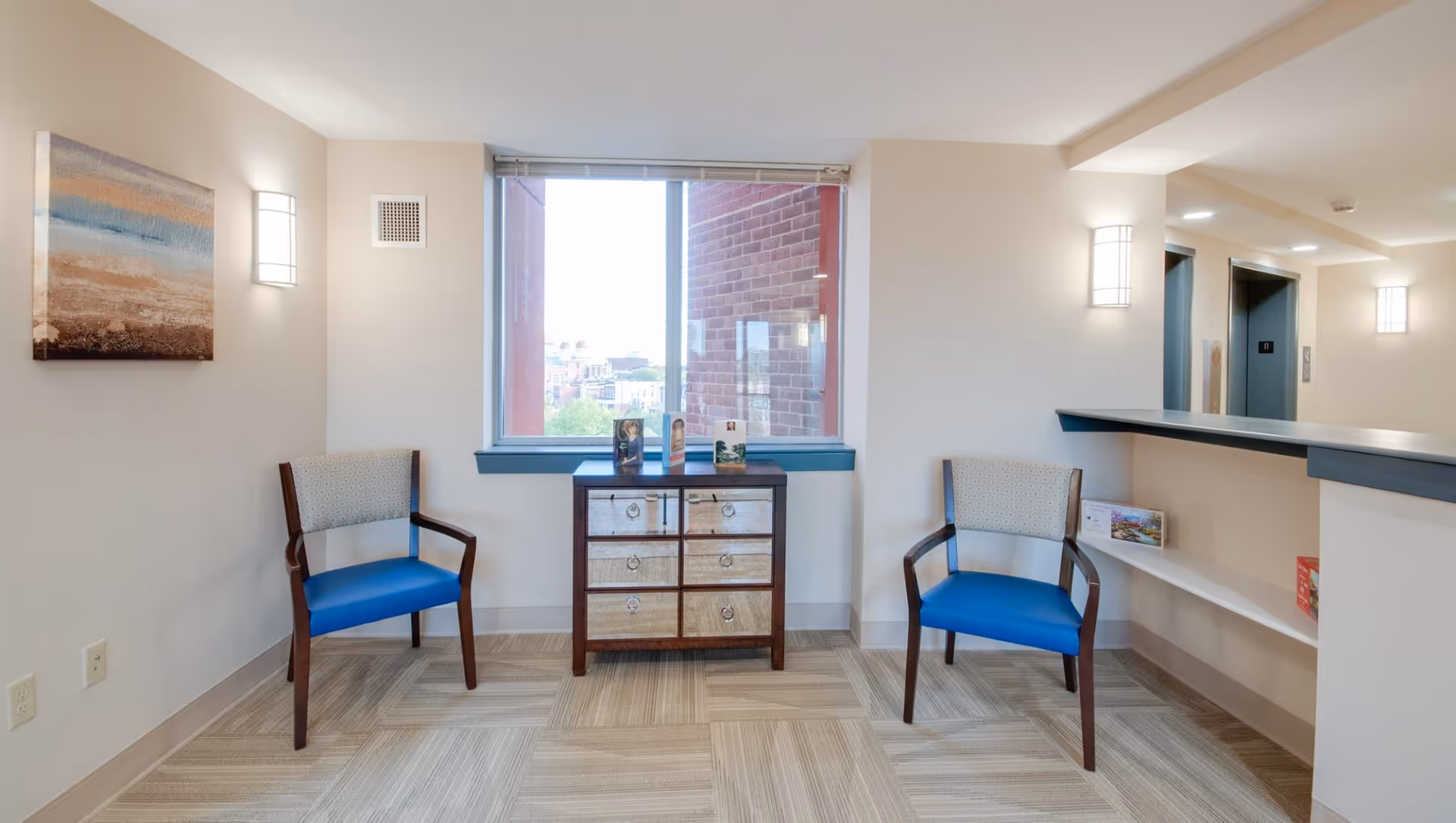 A small waiting area in a senior living facility with two wooden chairs with blue seats and patterned backs, a small wooden cabinet with six drawers, and a large window showing a partial outdoor view. The walls are light-colored with modern wall sconces providing lighting. There is a counter on the right side with a shelf underneath holding some books or brochures.