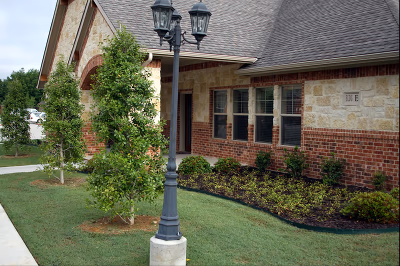 Exterior view of a building at Mustang Creek Estates Keller with a brick and stone facade, several windows, a small garden with shrubs and trees, and a black lamppost in the foreground.