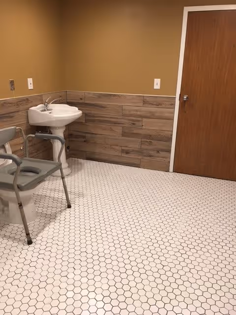 A bathroom with a white hexagonal tile floor, a white pedestal sink, a wooden door, and a gray commode chair positioned next to the sink. The walls are painted beige with a wooden panel halfway up.