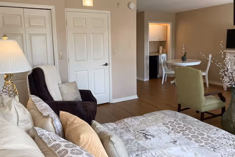 Sunlit studio apartment interior with a bed in the foreground, a recliner, small dining table, and a kitchenette area.