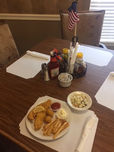 A plate of fried fish, french fries and sides on a dining table with placemats, condiments and a small American flag centerpiece.