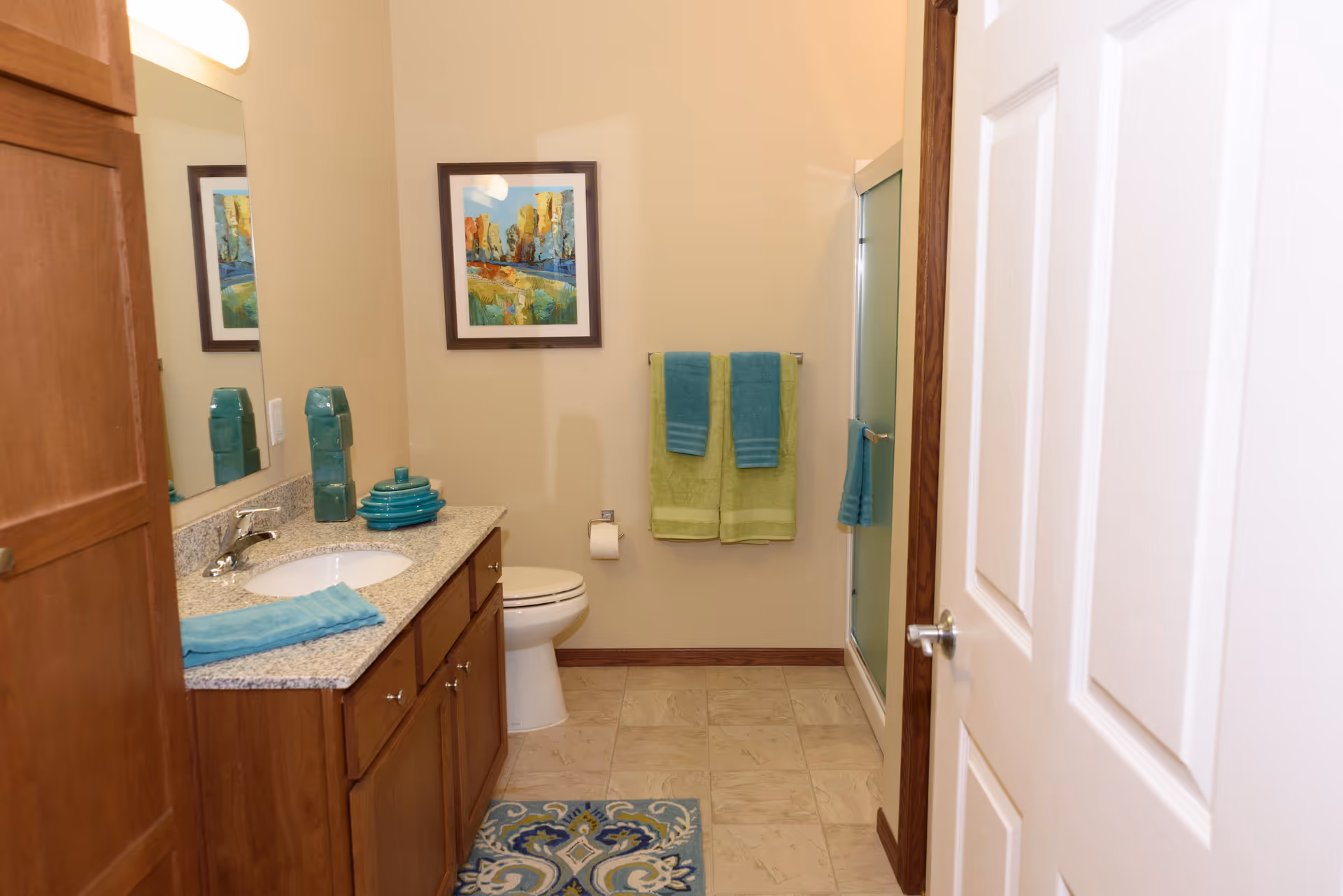 Bathroom with a wooden vanity and sink, toilet, glass shower, and teal/green towels and accessories.
