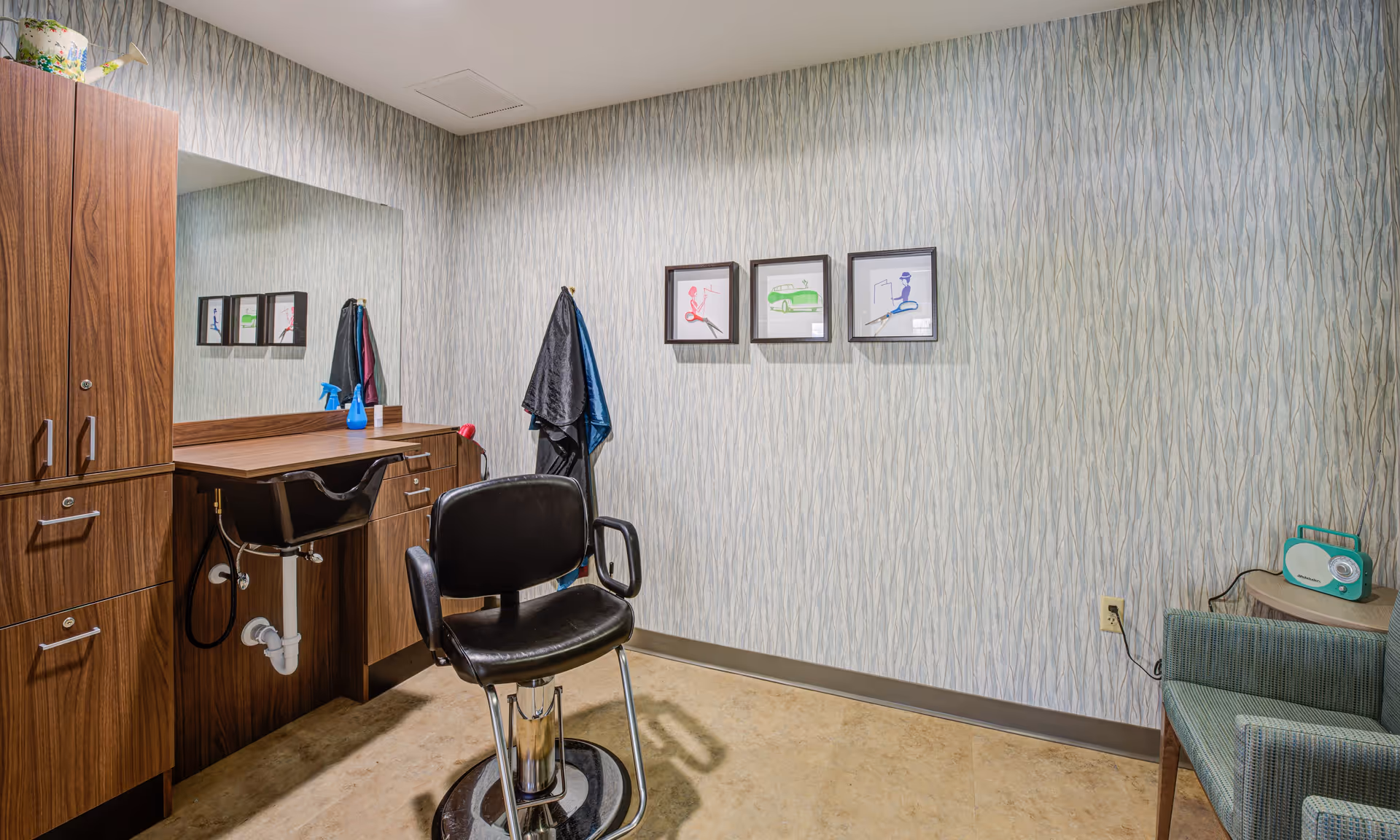 A small salon or grooming room with a black salon chair in front of a wooden counter and sink. There are cabinets on the left side, a large mirror above the counter, and three framed pictures on the wall. A coat rack with two capes hangs on the wall, and a green radio sits on a small table next to a green upholstered chair on the right.