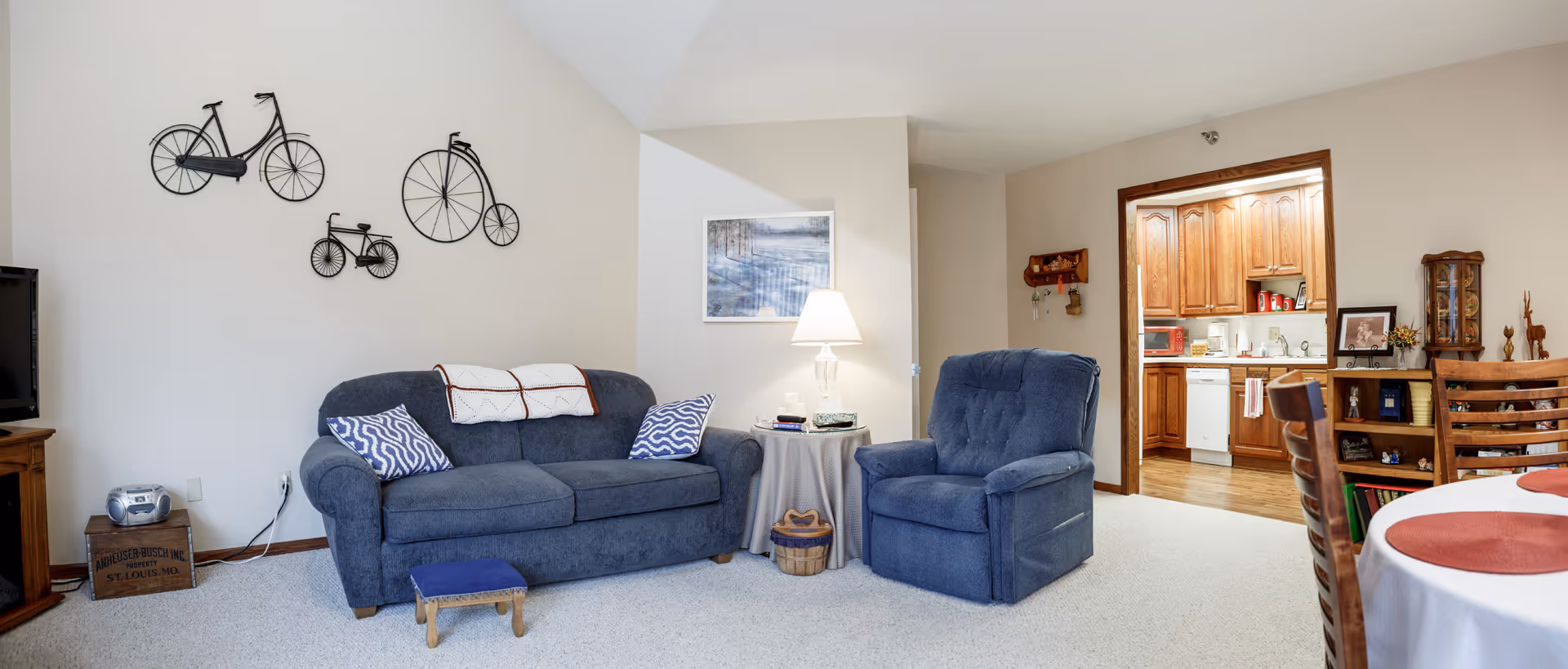 A cozy living room with a blue sofa and matching armchair, a small wooden footstool, and a round side table with a lamp and books. The wall behind the sofa features decorative metal bicycle art. To the right, there is a view into a kitchen with wooden cabinets and white appliances. A dining table with red placemats is partially visible in the foreground.