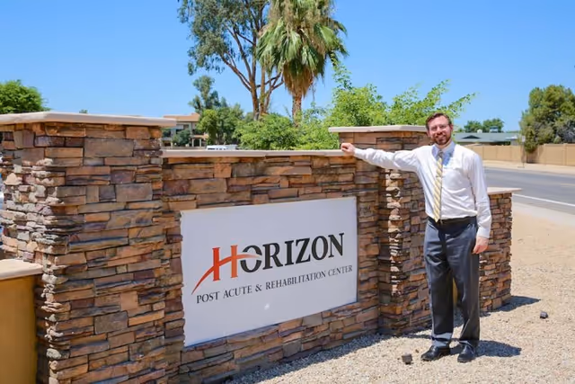 A man in business attire stands next to a stone sign for Horizon Post Acute & Rehabilitation Center on a sunny day with trees and a clear blue sky in the background.