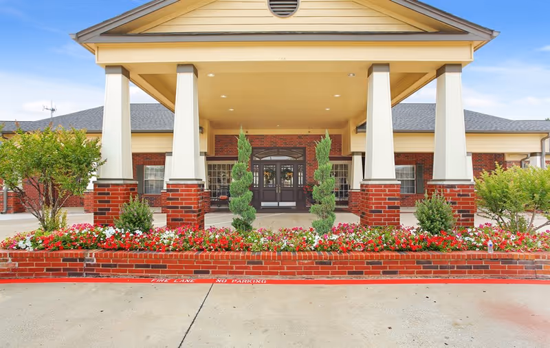 Front entrance of Bedford Estates Senior Living facility featuring a covered drop-off area with large white columns on brick bases, a flower bed with red and white flowers, and a clear blue sky in the background.