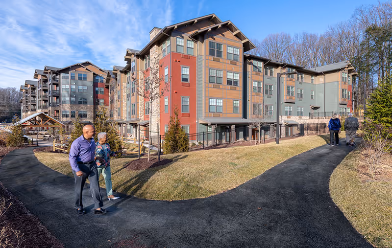A senior living facility with a multi-story building featuring a mix of red, brown, and green exterior panels. Four people are walking on paved paths surrounded by grass and landscaping. Trees without leaves are visible in the background under a blue sky with some clouds.