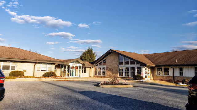 Exterior view of a single-story senior living facility building with a brown roof and large windows under a blue sky with some clouds. There is a paved parking area in front with a few parked cars and some small bushes near the building.