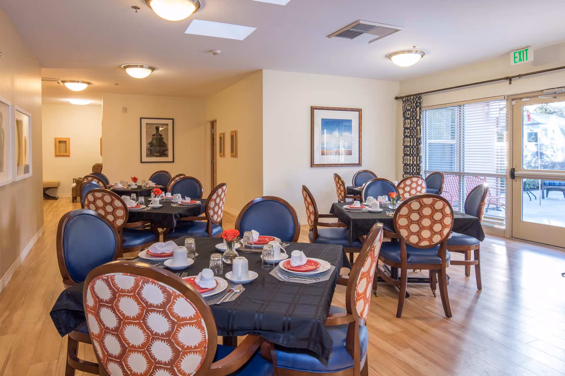 A bright dining room with several round tables covered in black tablecloths, each set with white cups, plates, and red napkins. The chairs have blue seats and backs, with some featuring a patterned fabric on the backrest. Large windows and a glass door let in natural light, and framed artwork hangs on the walls.