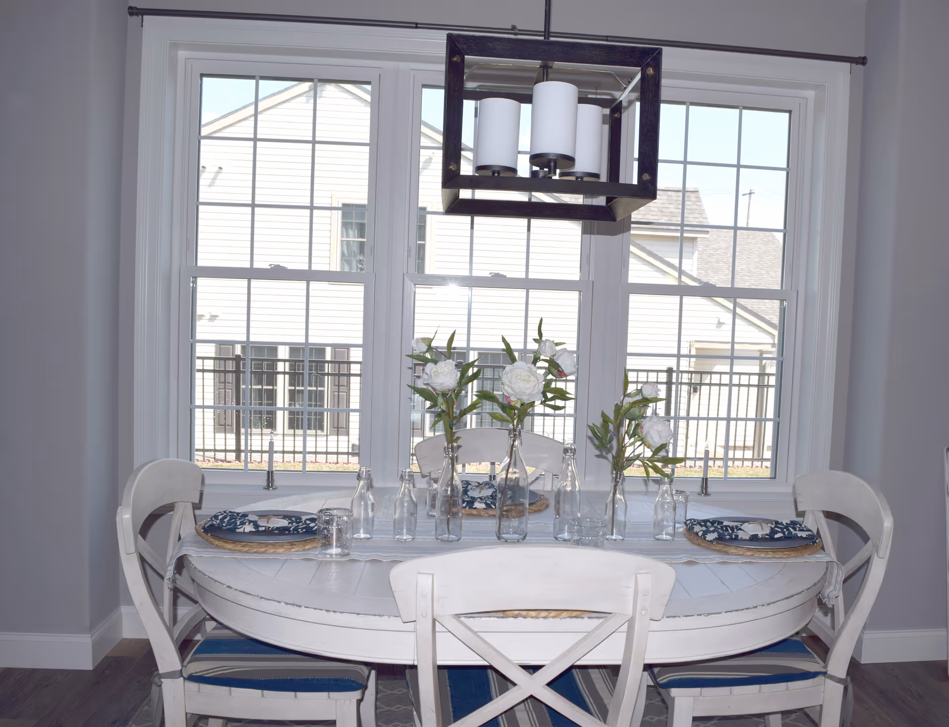 Dining area with a round white table set for four, glass vases with white flowers, and a large window with a hanging light fixture.