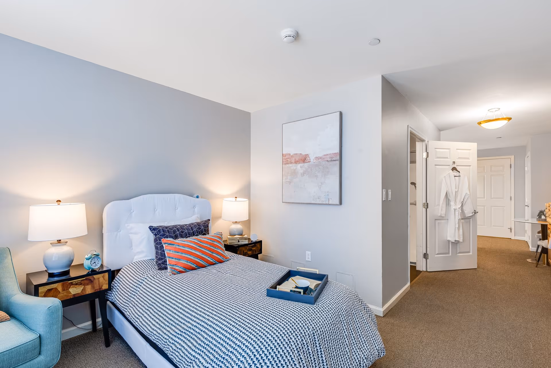 A neatly arranged bedroom with a single bed featuring a white tufted headboard, blue and orange striped pillows, and a blue and white patterned blanket. Two bedside tables with matching lamps flank the bed. A light blue armchair is positioned to the left. A white robe hangs on the open door leading to a hallway with beige carpet and white walls. A piece of abstract art is mounted on the wall above the bed.
