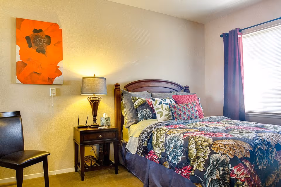 A brightly lit bedroom with a floral-patterned bed, wooden headboard, nightstand with lamp, and colorful wall art.
