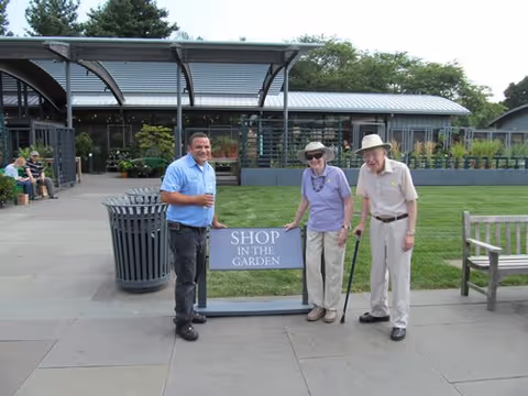 Three people standing outdoors near a sign that reads 'SHOP IN THE GARDEN'. Two elderly individuals, one with a cane, are standing next to a younger man holding a drink. They are in a garden area with a building and greenery in the background.