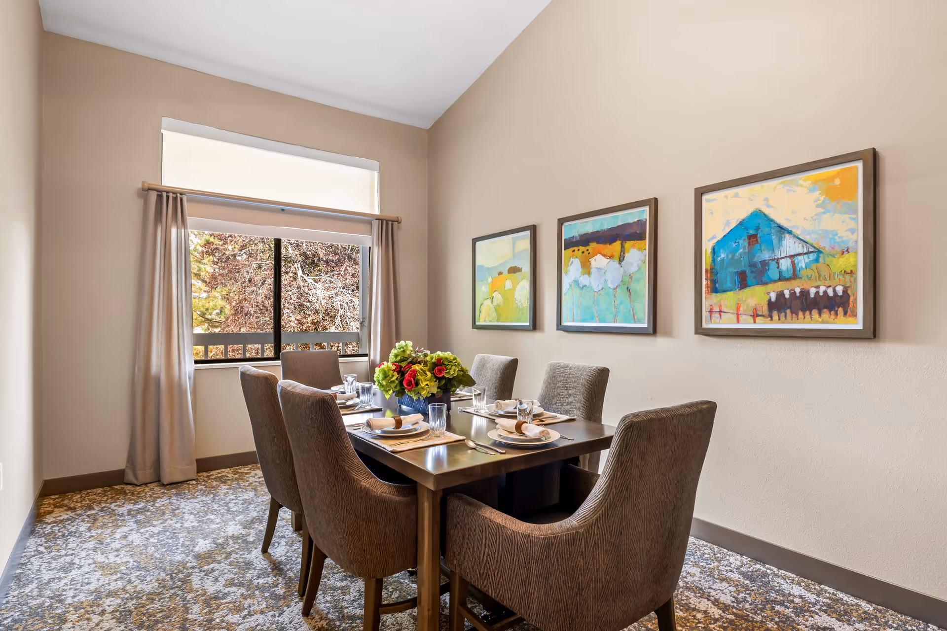 A dining room with a rectangular wooden table set for six people with plates, glasses, and napkins. There is a floral centerpiece on the table. The room has beige walls with three colorful paintings hanging on one wall. A window with beige curtains allows natural light to enter, and the floor is covered with a patterned carpet.