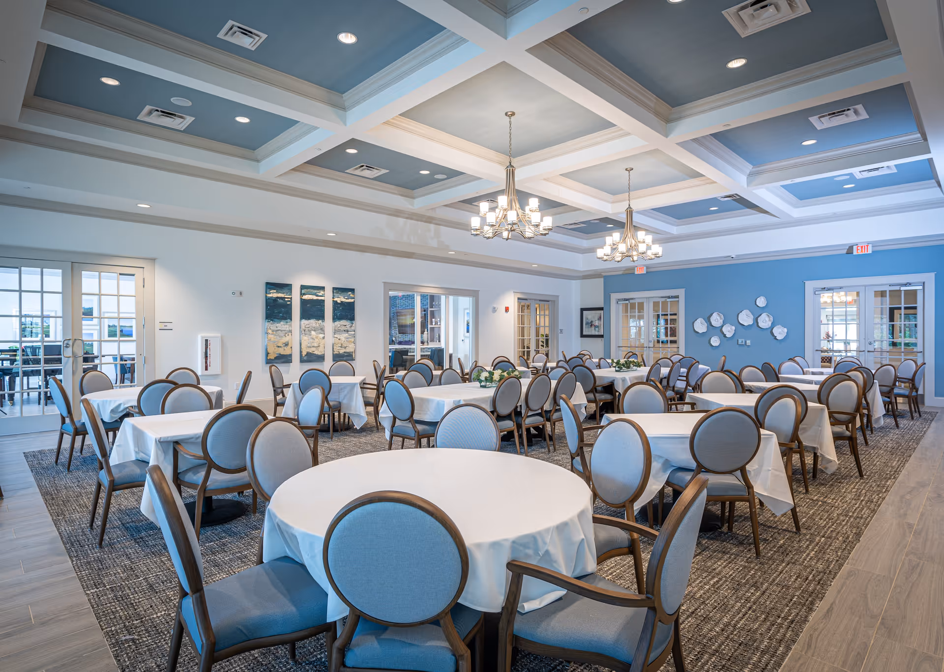 A spacious dining room with multiple round and rectangular tables covered with white tablecloths, surrounded by wooden chairs with blue cushions. The room features a coffered ceiling painted blue with recessed lighting and two chandeliers. The walls are white and blue with decorative artwork and floral arrangements on some tables. There are glass doors leading to other rooms and an outdoor area.