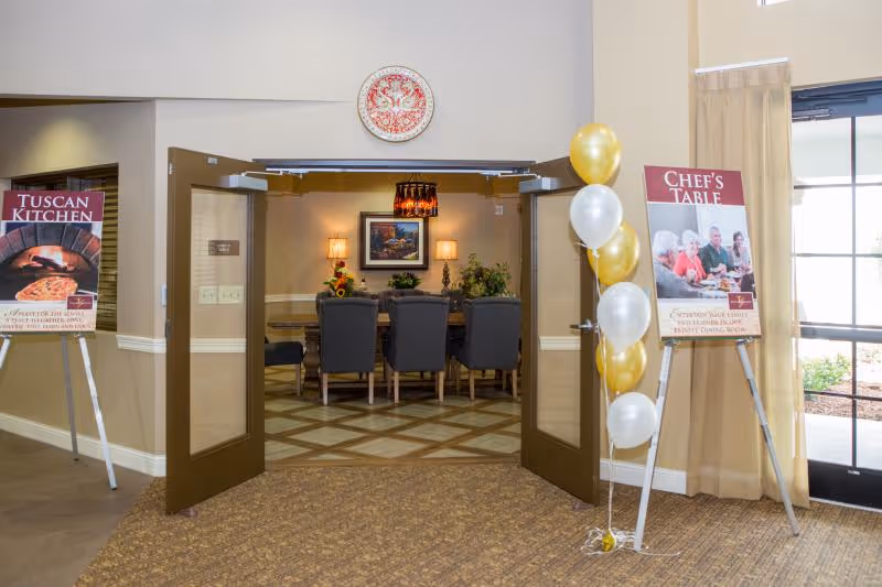 Open double doors reveal a formal dining room with a long table and chairs, flanked by a 'Tuscan Kitchen' sign on the left and a 'Chef's Table' sign with gold and white balloons on the right.