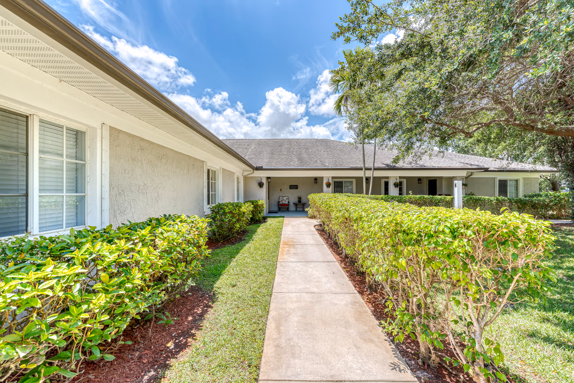 A sunny exterior view of a single-story building with a concrete walkway leading to a covered entrance. The building is surrounded by neatly trimmed green hedges and trees under a bright blue sky with scattered clouds.