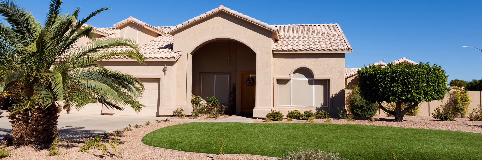 Single-story beige stucco house with a tiled roof, a large arched entryway, and a garage. The front yard features a well-maintained green lawn, a palm tree on the left, and a neatly trimmed round tree on the right under a clear blue sky.