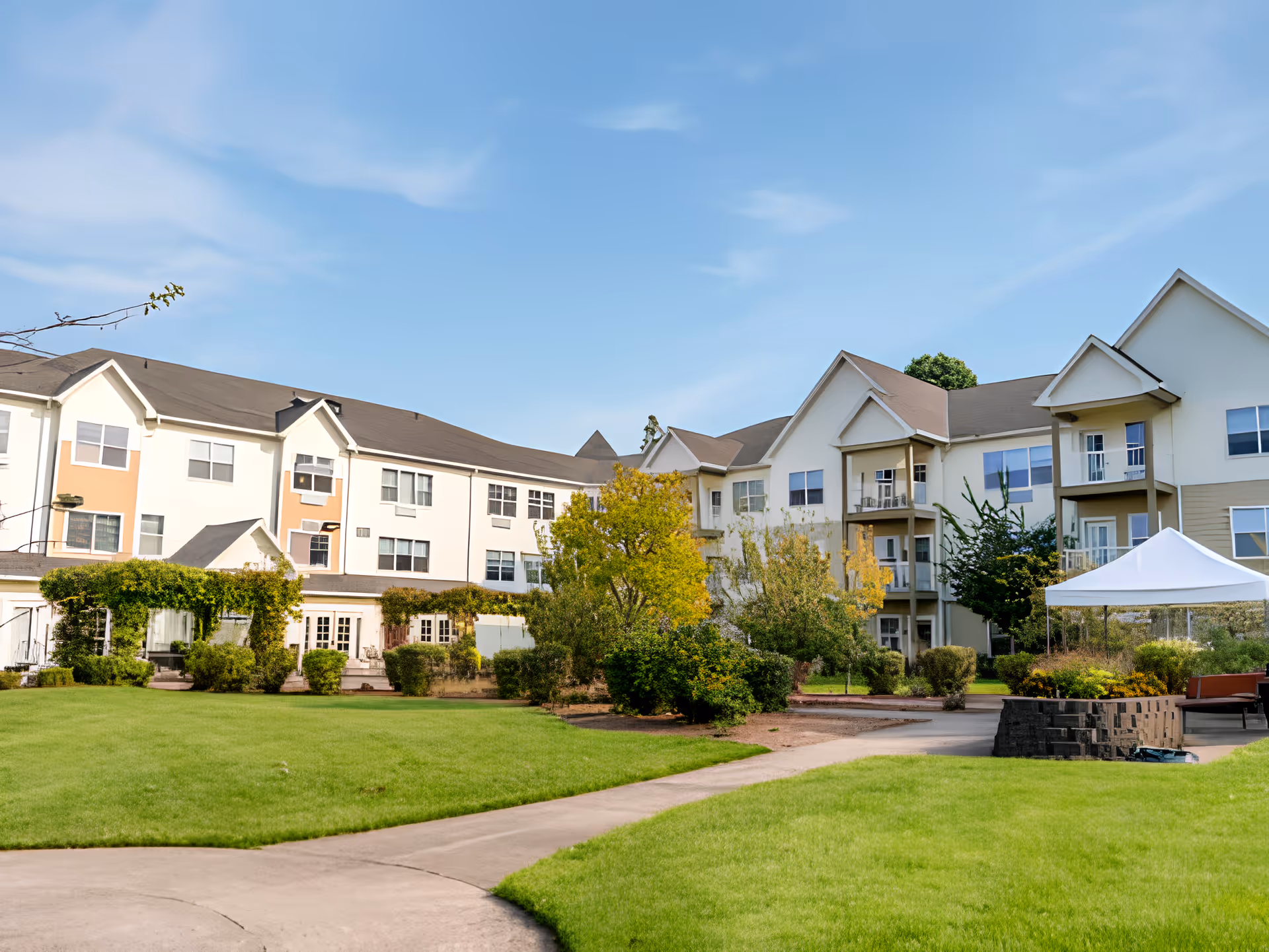 Exterior view of a multi-story senior living facility building with beige and white walls, surrounded by green lawns, trees, and shrubs under a blue sky. A paved walkway curves through the lawn leading to the building, and a white canopy tent is set up near the right side.