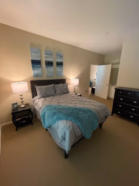 A neatly made bed with gray bedding and a teal throw blanket in a bedroom. Two bedside tables with lamps flank the bed. Above the bed are three vertical abstract paintings in shades of blue and white. To the right, there is a black dresser with multiple drawers. An open door reveals a glimpse of an adjoining room with a sink and mirror.