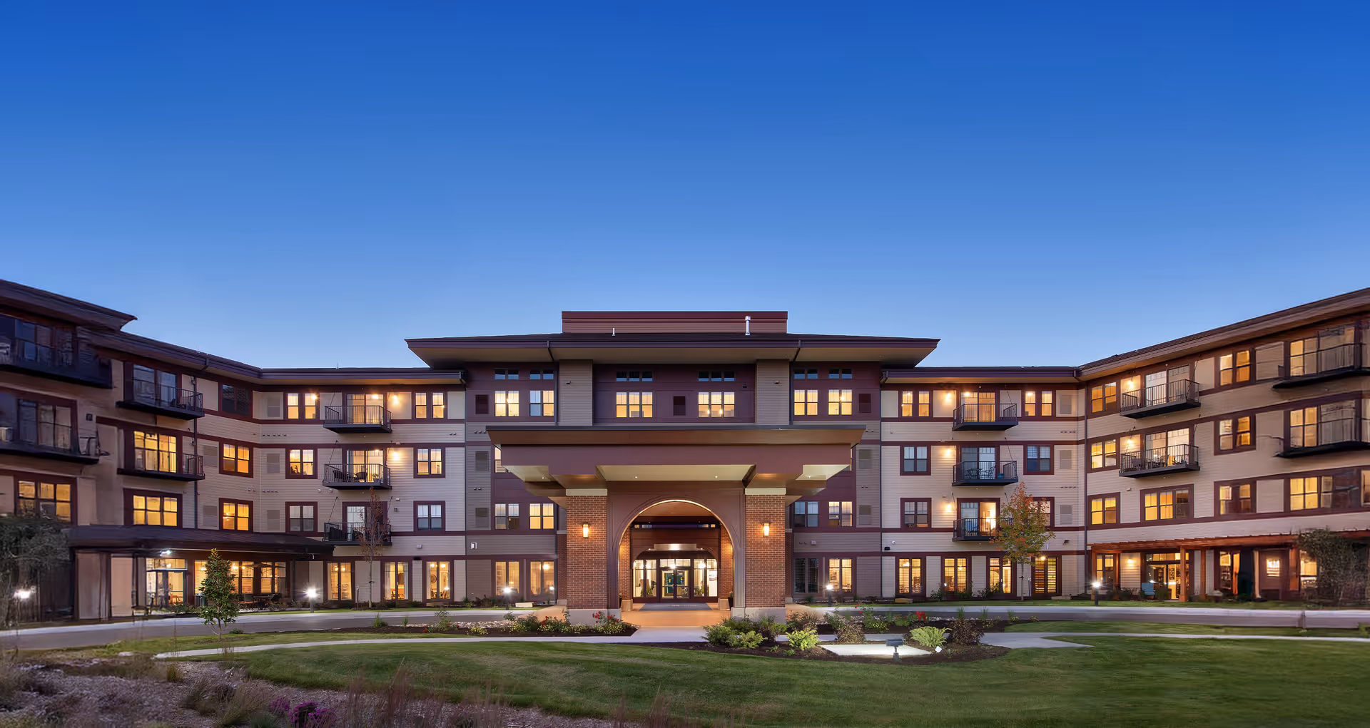 Exterior view of a large, modern senior living facility building at dusk with multiple windows lit and a covered entrance in the center. The building is surrounded by a well-maintained lawn and landscaped garden areas under a clear blue sky.