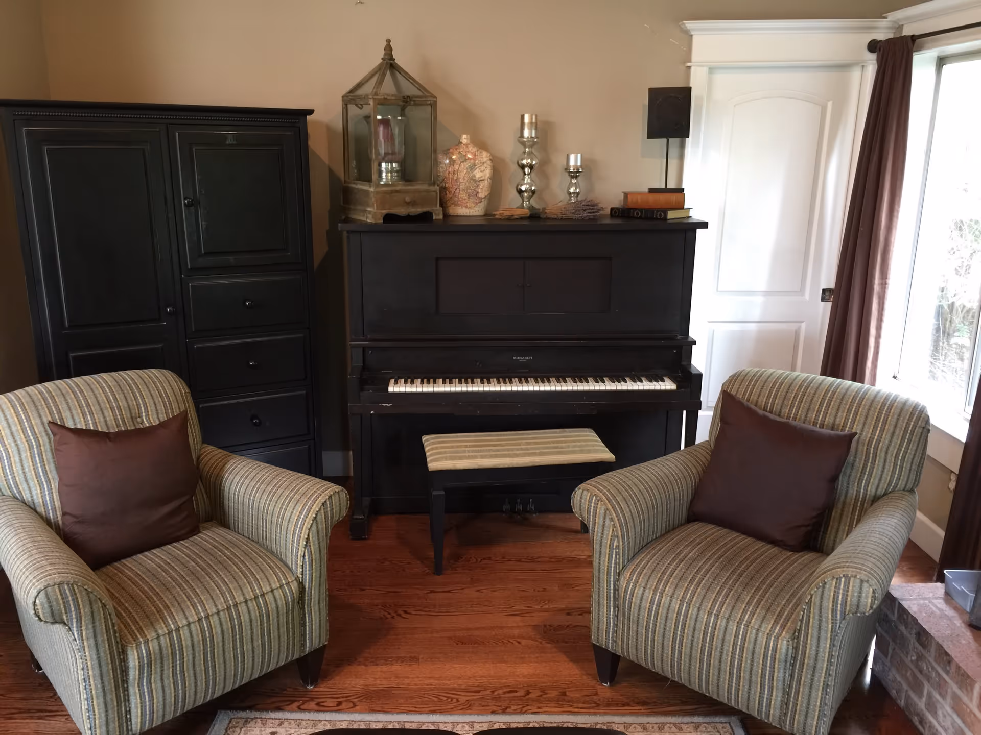 A cozy living room area with two striped armchairs each having a brown cushion, a black upright piano with a matching bench, and a black cabinet. On top of the piano are decorative items including a lantern, a vase, candles, and some books. There is a white door and a window with brown curtains on the right side.