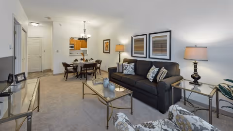Carpeted living room and dining area featuring a dark sofa, glass-top coffee table, armchair, side tables with lamps, and a small dining table near a kitchen pass-through.