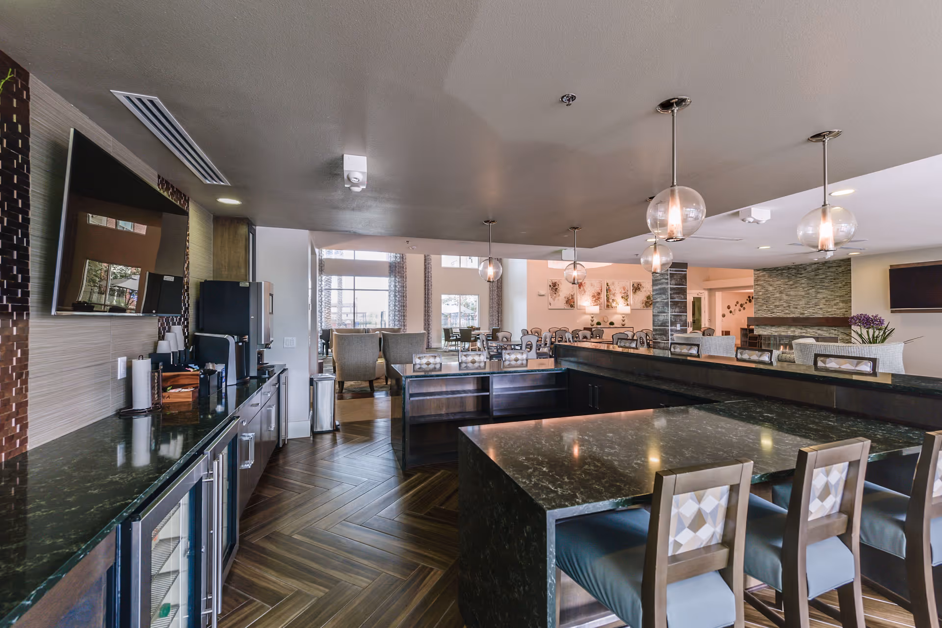 Modern communal dining and lounge area with a large dark marble countertop island surrounded by chairs, pendant lights hanging from the ceiling, and a seating area with tables and chairs in the background. The space features wood-patterned flooring and large windows allowing natural light.