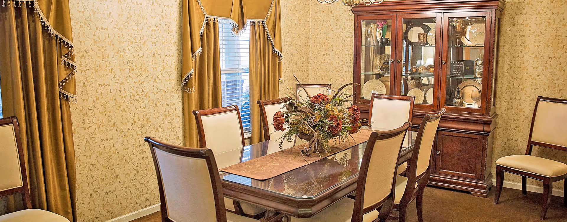 A formal dining room with a rectangular wooden table surrounded by six upholstered chairs. The table has a decorative centerpiece with flowers and foliage. Behind the table is a wooden china cabinet displaying plates and decorative items. The walls are covered with patterned wallpaper, and there are gold-colored curtains framing a window.