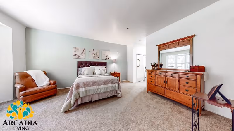 A spacious bedroom with a bed centered against a light blue accent wall, flanked by a wooden nightstand with a lamp. To the left, there is a brown leather armchair with a white throw blanket. On the right side, a large wooden dresser with a mirror and decorative items is visible. The room has beige carpet flooring and white walls, with a doorway leading to another area.