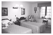 A bedroom with two twin beds, each with a quilted bedspread and stuffed animals placed on top. The room has a window with patterned curtains, a small table beneath the window, framed pictures on the walls, and a wall-mounted light fixture above the beds.