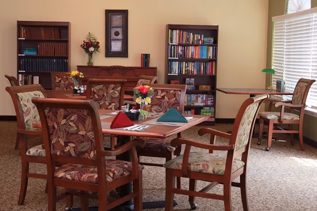 Dining room with wooden tables and upholstered chairs set with folded napkins and small flower arrangements, with bookshelves and a piano against the wall.