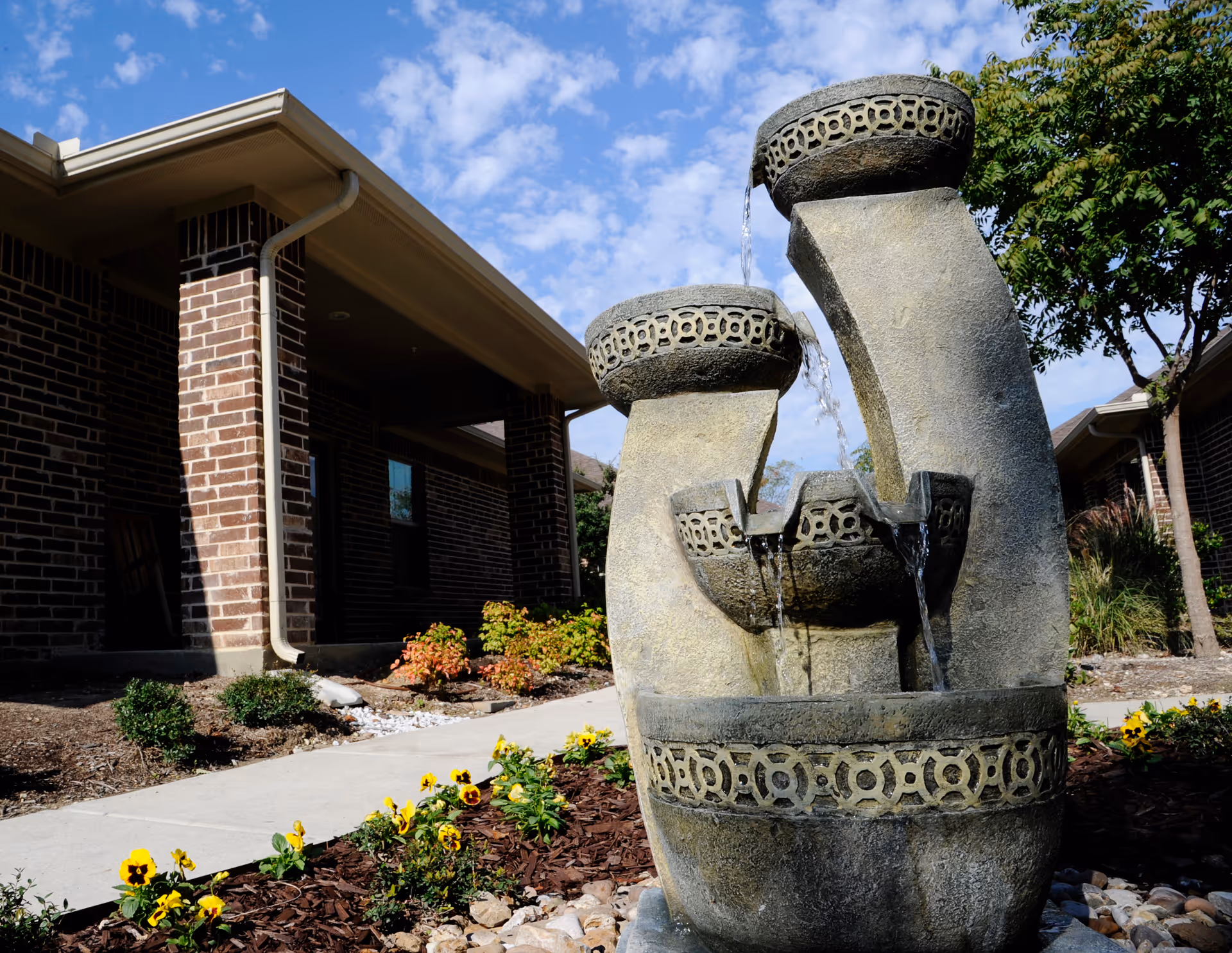 Outdoor scene at Mayberry Gardens Assisted Living and Memory Care Homes featuring a decorative stone water fountain with water flowing from three tiers. The fountain is surrounded by a landscaped garden with yellow flowers and mulch. In the background, there is a brick building with a covered walkway and trees under a partly cloudy blue sky.