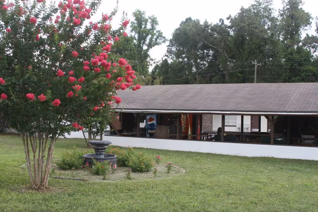 Outdoor view of a grassy area with a flowering tree and a small circular garden with a fountain in the center. In the background, there is a long building with a covered porch area that has vending machines visible inside.