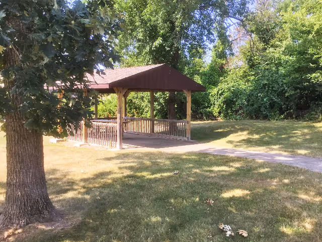 A wooden gazebo on a grassy lawn surrounded by trees and a paved walkway.