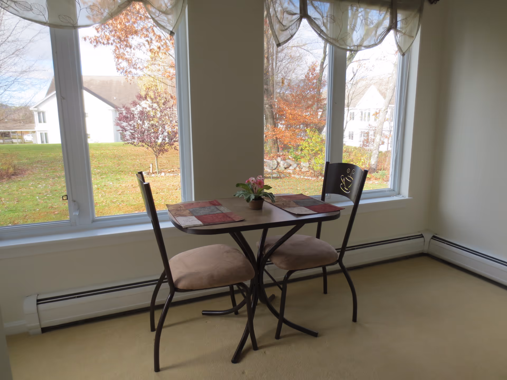 A small dining area with a square table and two chairs with cushioned seats. The table has two placemats and a small potted plant in the center. Large windows behind the table show a view of a grassy yard with trees and neighboring houses.