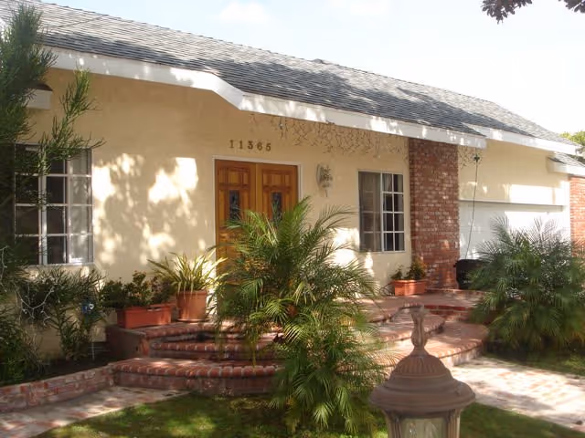 Front exterior view of a single-story building with a beige facade, brick accents, and a shingled roof. There are two wooden doors in the center with the number 11365 above them. The entrance is surrounded by potted plants and small palm trees, with a brick pathway and steps leading up to the doors. A decorative lamp post is visible in the foreground.