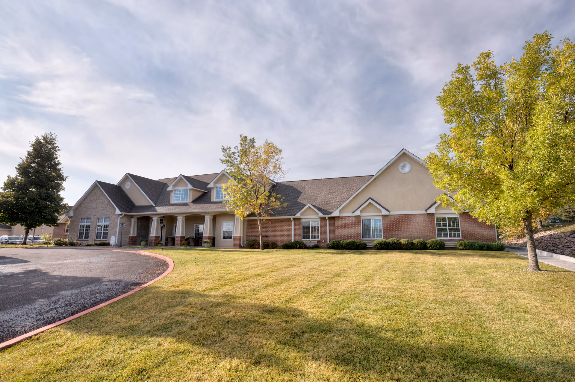 Exterior front view of a single-story brick and stucco senior living building with a lawn, trees, and a curved driveway.