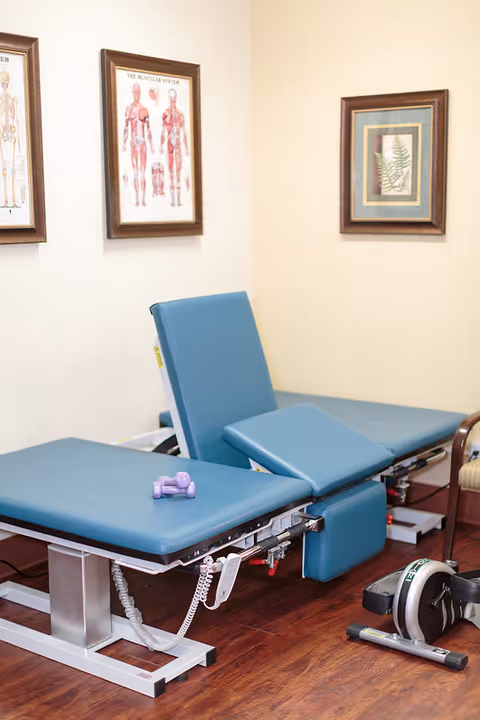 A therapy or examination room with two blue padded adjustable treatment tables, a small purple dumbbell on one table, and a compact exercise bike on the wooden floor. The walls have framed anatomical and botanical pictures.