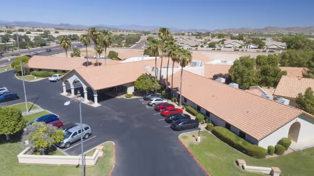 Aerial view of Lake Pleasant Post Acute Rehabilitation Center showing a single-story building with a tiled roof, surrounded by a parking lot with several cars and landscaped greenery including palm trees. Residential neighborhoods and mountains are visible in the background under a clear blue sky.