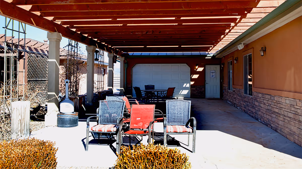 Outdoor covered patio area with several chairs and tables arranged for seating. The patio has a wooden pergola roof supported by columns, and the surrounding area includes a building wall with windows and a door, as well as some landscaping with bushes and a fence.