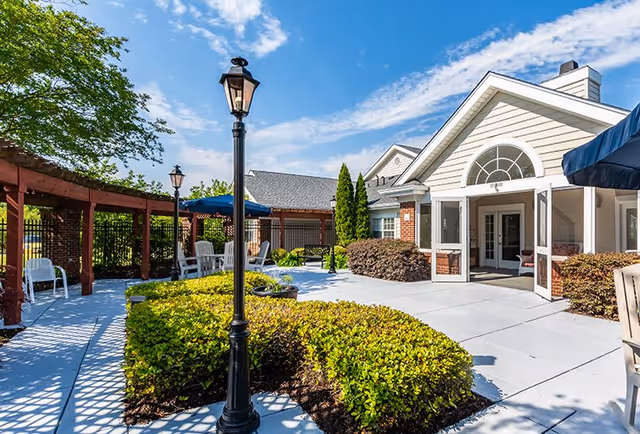 Outdoor patio area at Legacy Heights Senior Living Community featuring a paved walkway, black lamp posts, green bushes, white chairs under a wooden pergola, and a building entrance with large windows and a glass door under a blue sky with scattered clouds.