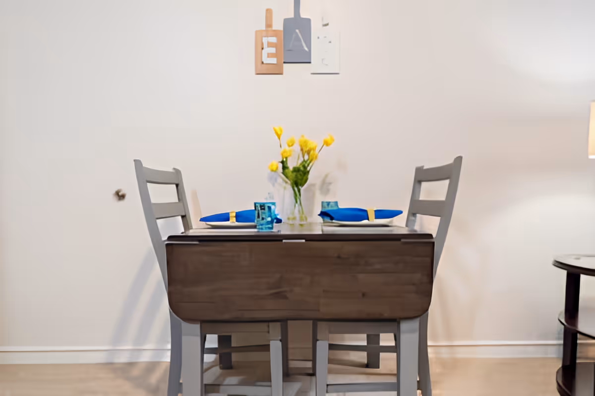 A small dining table set for two with blue napkins and glasses, a vase with yellow flowers in the center, two gray chairs, a floor lamp to the right, and three decorative wall hangings above the table.